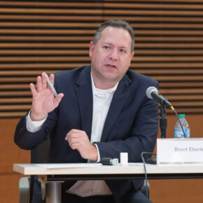 Brent Eberle speaking into a microphone while seated at a table and gesturing with his hand