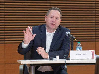Brent Eberle speaking into a microphone while seated at a table and gesturing with his hand