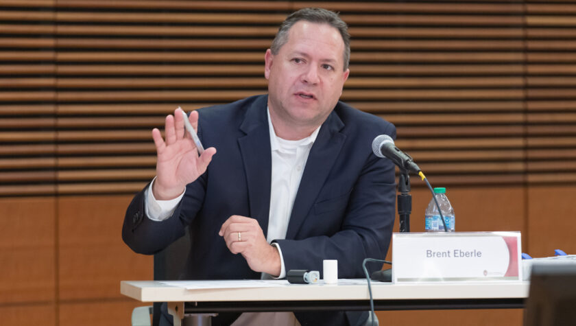 Brent Eberle speaking into a microphone while seated at a table and gesturing with his hand