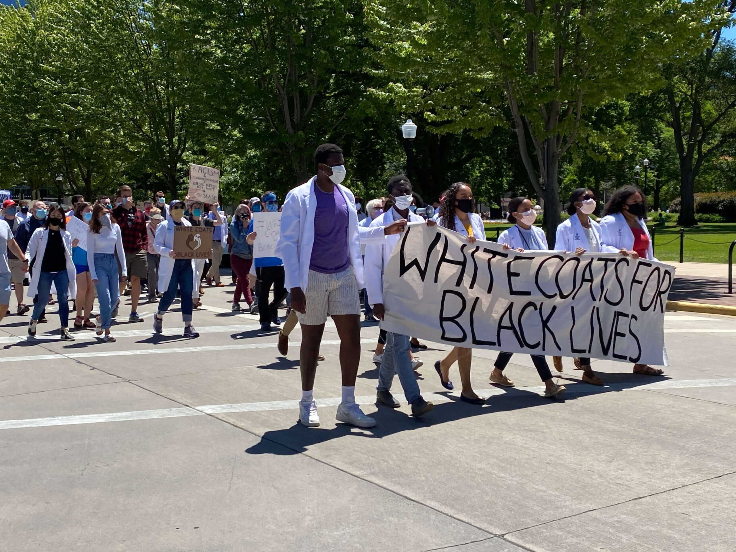 Pharm students wearing white coats and masks walking in protest with a sign reading "White Coats for Black Lives"