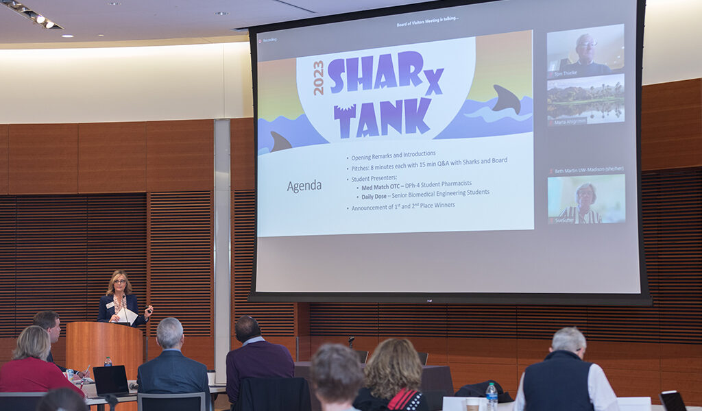 Beth Martin speaks in front of a large screen bearing the SHARx Tank logo
