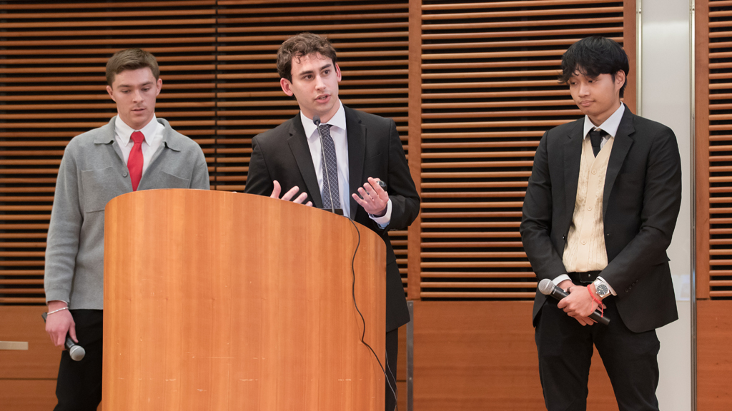 Three PharmD students in jackets and ties stand near a lectern. The one in the middle is speaking into a microphone