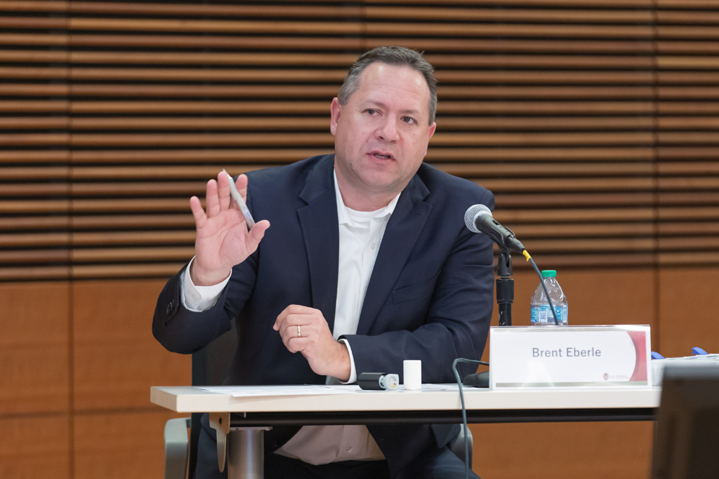 Brent Eberle speaking into a microphone while seated at a table and gesturing with his hand