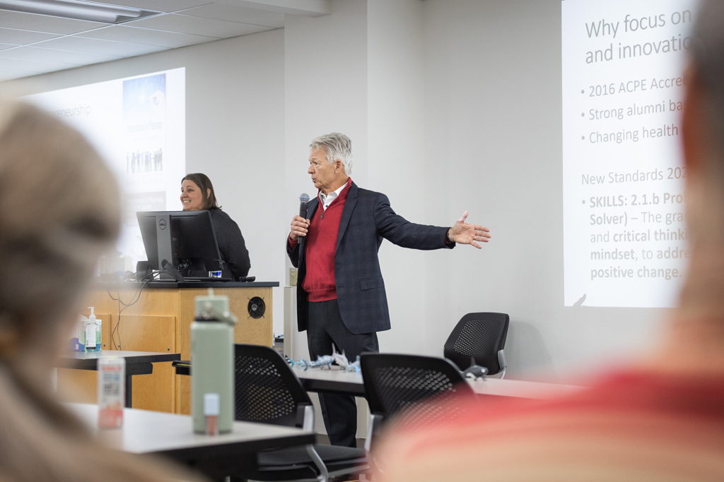 George Zorich gestures in front of a projected screen while speaking into a microphone