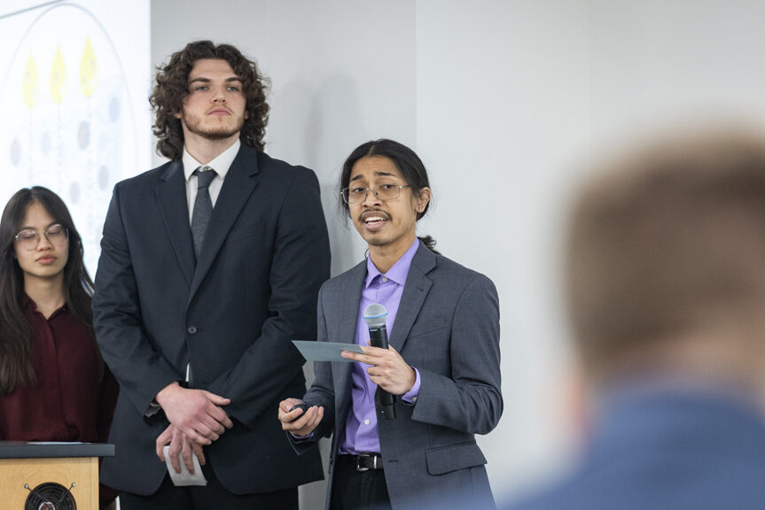 SharX Tank 2026 A PharmD student in a purple shirt and gray jacket speaks into a microphone in front of his team