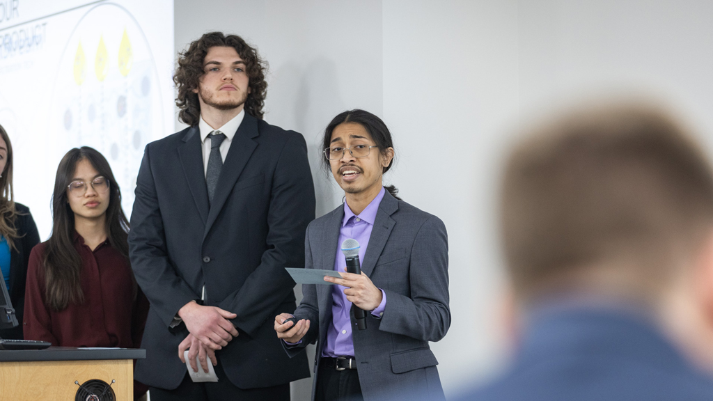 A PharmD student in a purple shirt and gray jacket speaks into a microphone in front of his team