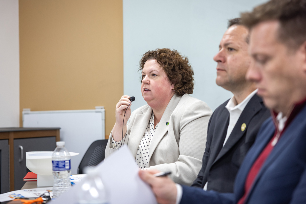 Sarah Sorum speaks into a small microphone while seated at a table alongside Brent Eberle and Joel Jones