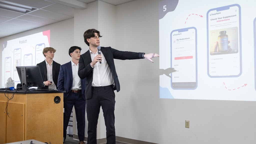 A PharmD student holding a microphone points toward a projection on a wall showing a cellphone app