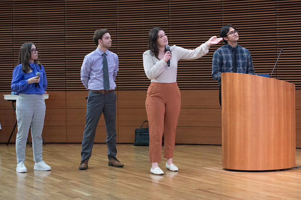 PharmD students Maite Zapata, Alexander Weida, Jenna Vande Hey, and Theo Buna stand in a line, with Jenna holding a microphone, talking, and gesturing to the large screen