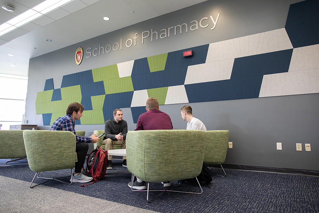 Comfortable lounge-style seating in front of a blue-and-green tiled wall with the "School of Pharmacy" logo on it. Students study in the seats.