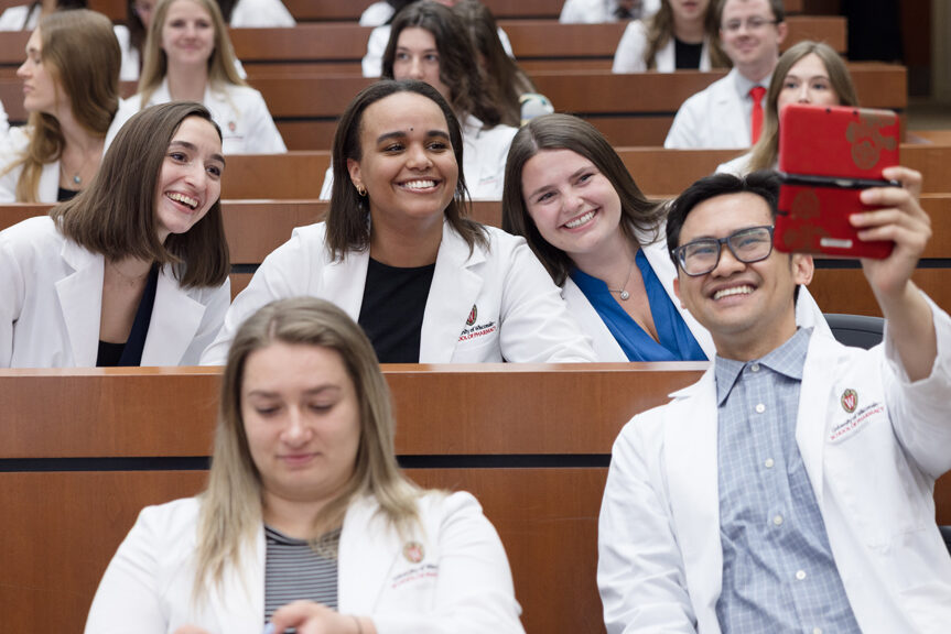 A group of PharmD students take a selfie in their white coats in a lecture hall