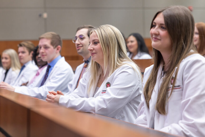 PharmD students in their white coats smile while seated in a a lecture hall