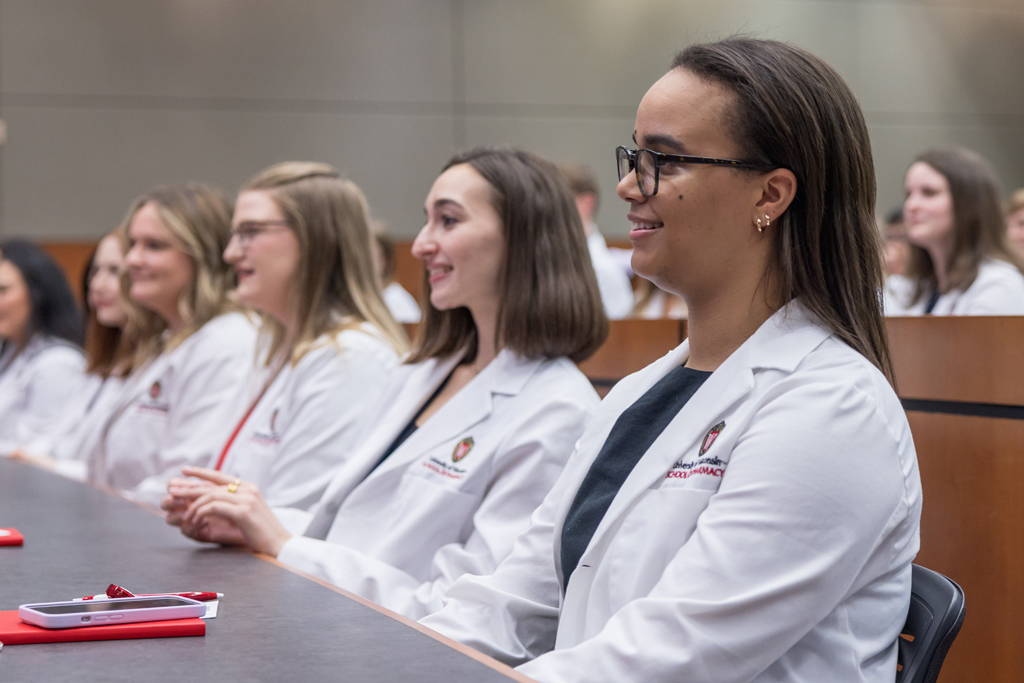 PharmD students in their white coats smile while seated in a a lecture hall