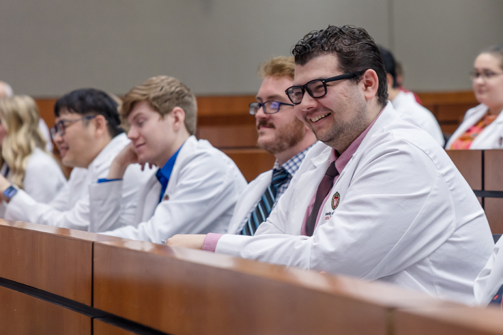 PharmD students in their white coats smile while seated in a a lecture hall