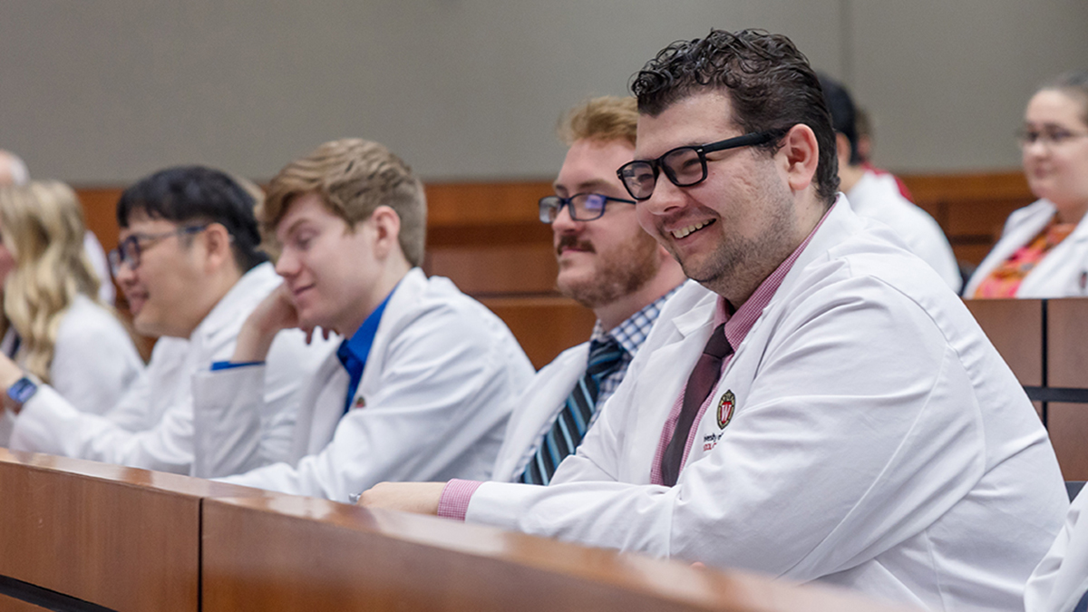PharmD students in white coats sit in a lecture hall and smile at a presenter