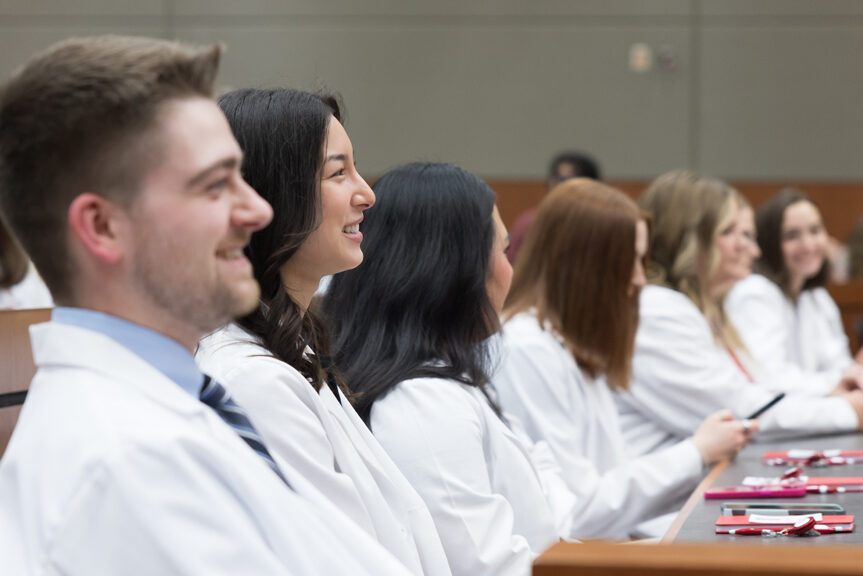PharmD students in their white coats smile while seated in a a lecture hall