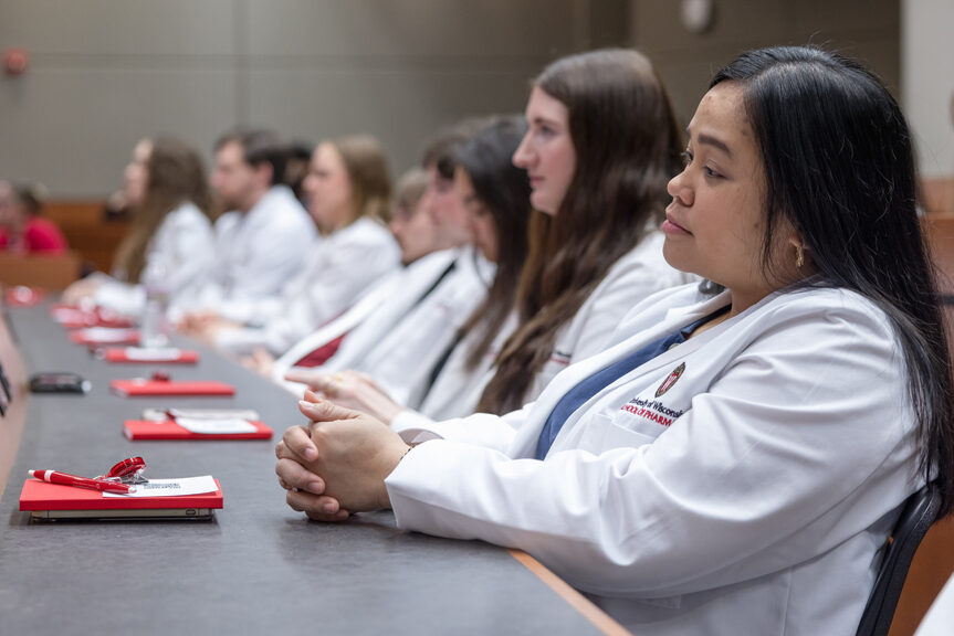 PharmD students in their white coats smile while seated in a a lecture hall