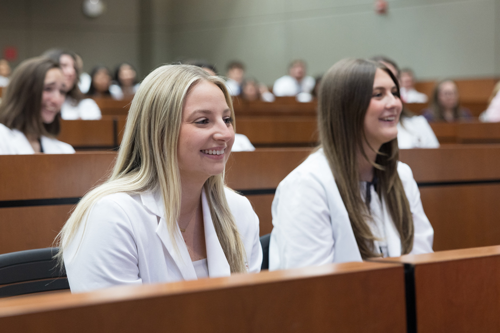 PharmD students in their white coats smile while seated in a a lecture hall