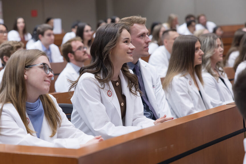 Seated PharmD students in white coats smile