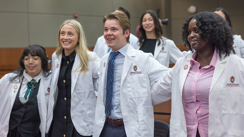 PharmD students in white coats stand arm in arm while smiling