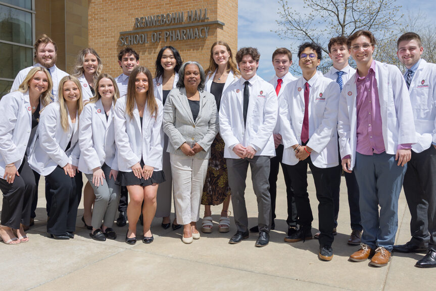 A large group of PharmD students pose with Eva Vivian in front of Rennebohm Hall in their white coats