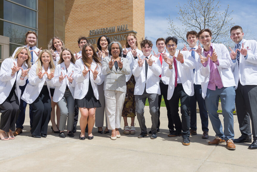 A group of PharmD students in white coats pose in front of Rennebohm Hall with W hands up