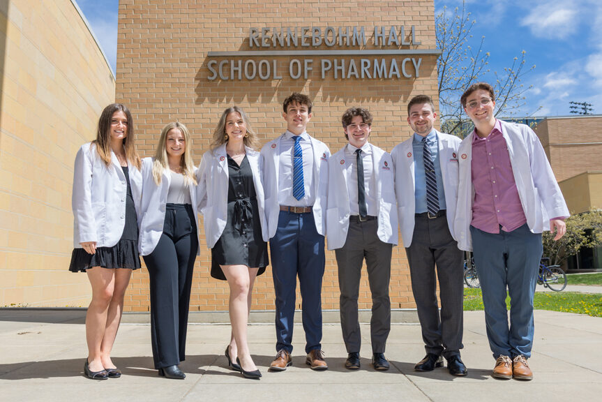 A group of PharmD students pose in front of Rennebohm Hall in their white coats