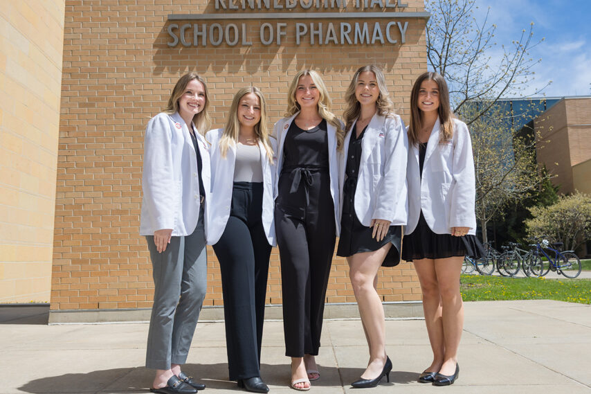 A group of PharmD students pose in front of Rennebohm Hall in their white coats