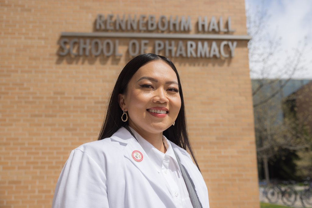 A PharmD student in her white coat posing in front of the Rennebohm Hall sign