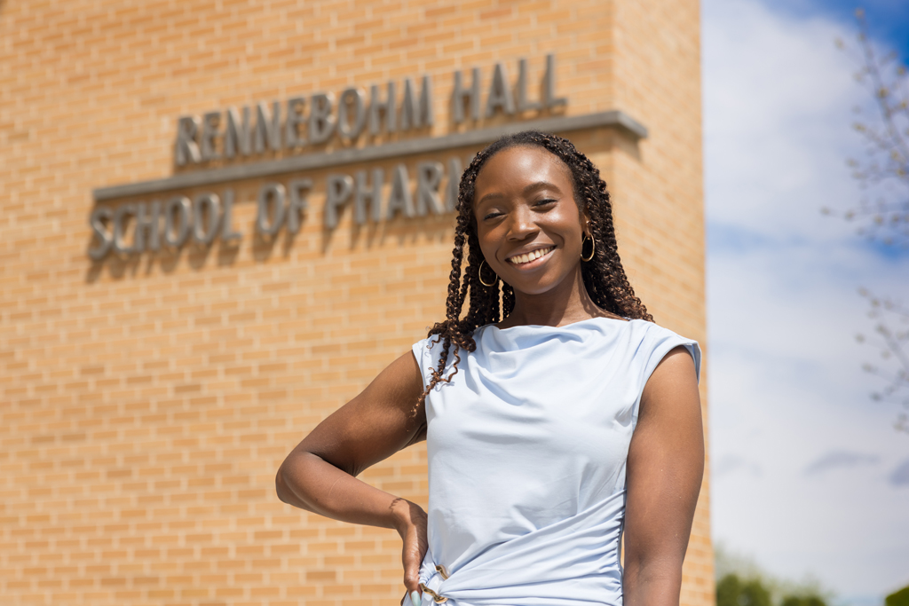 A PharmD student in a light blue dress posing in front of the Rennebohm Hall sign