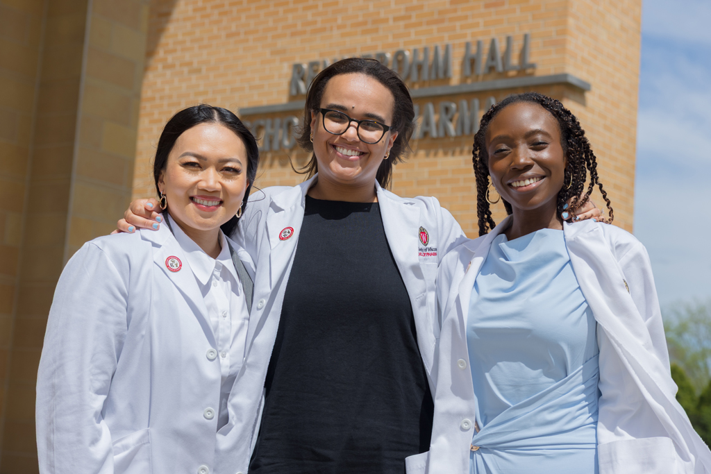 Three PharmD students in white coats posing in front of the Rennebohm Hall sign