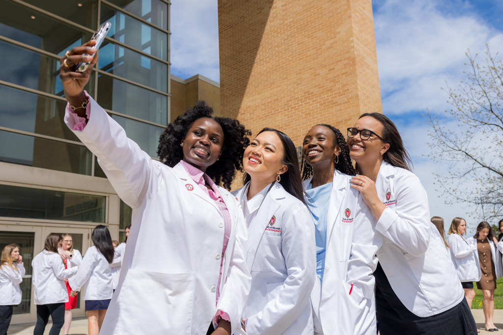 Four PharmD students in white coats taking a selfie outside Rennebohm Hall