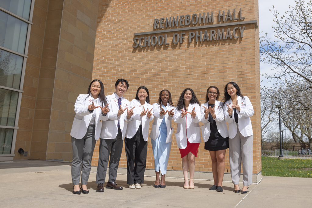 A group of PharmD students in white coats hold up W hands in front of the Rennebohm Hall sign