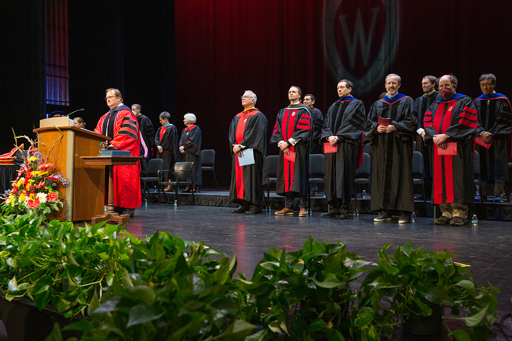 Pan out shot of professors standing onstage with Steve Swanson at the podium and hoods laying on tables to the left