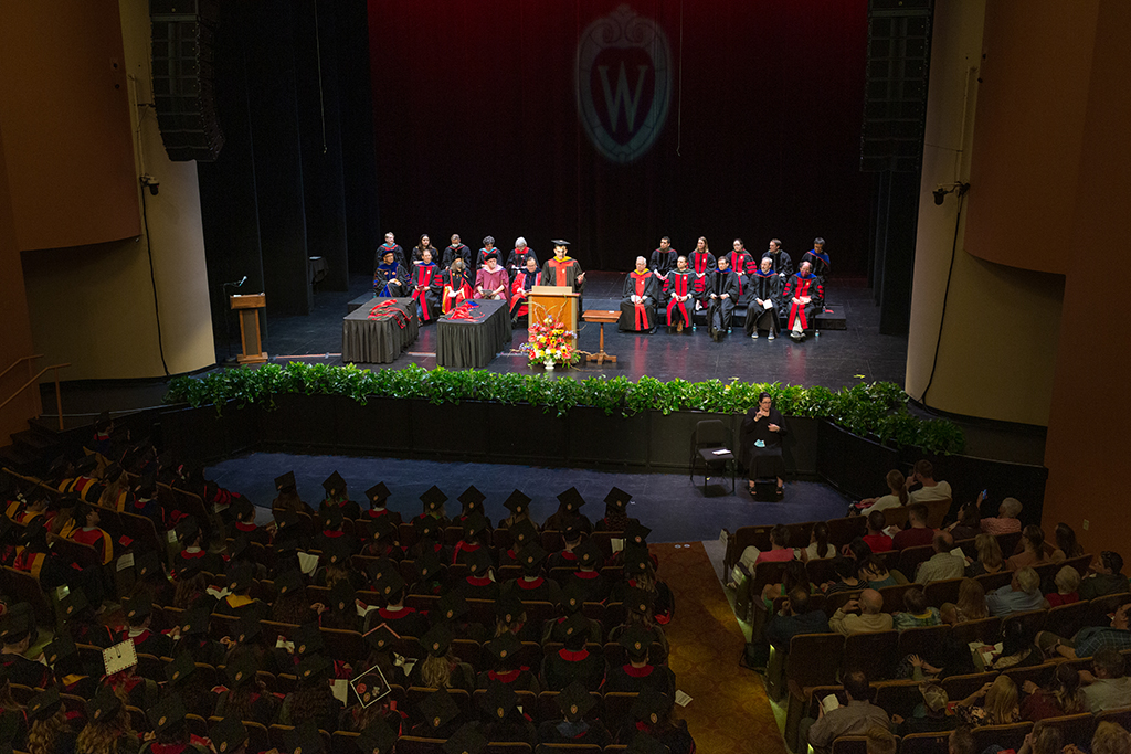 Pan out shot of professors sitting onstage with a speaker at the podium and hoods laying on tables to the left