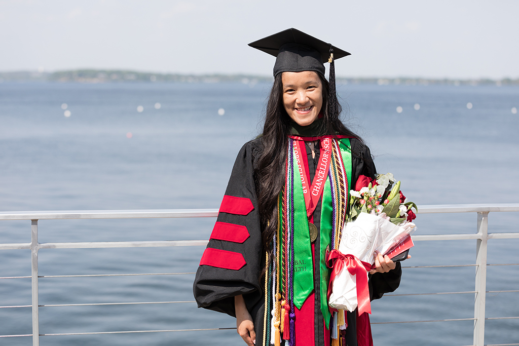 PharmD Class of 2022 graduate Stephaney Cheng poses in her cap and gown with a bouquet of flowers in front of Lake Mendota following the Hooding Ceremony