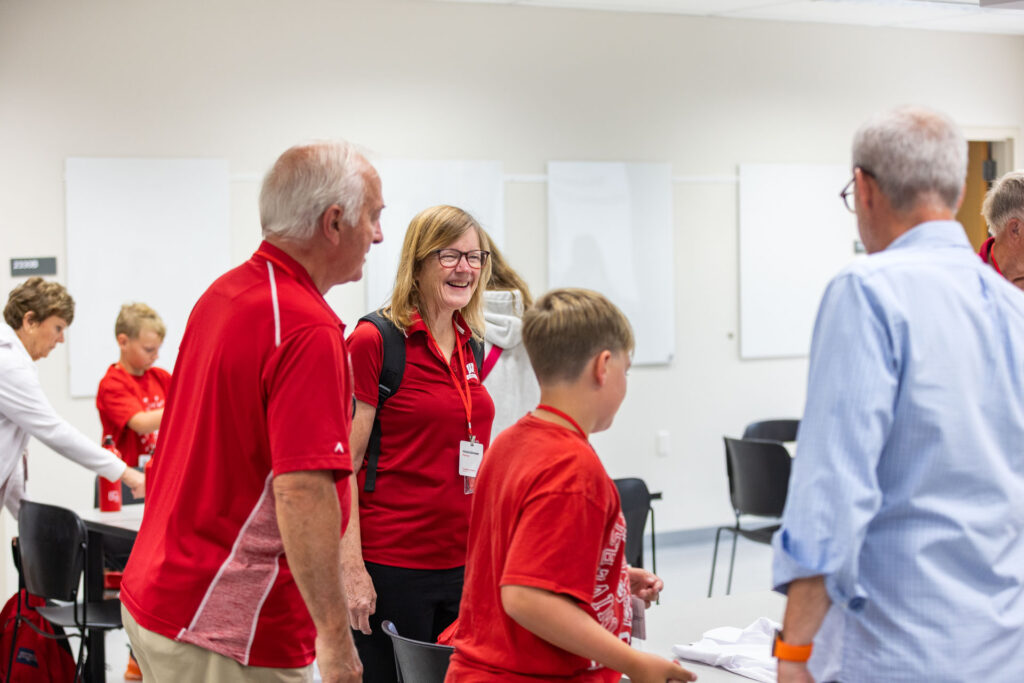 A group of smiling adults and children in red shirts are gathered, talking and laughing in a bright, modern room. An older man in a light blue shirt is partially visible on the right.
