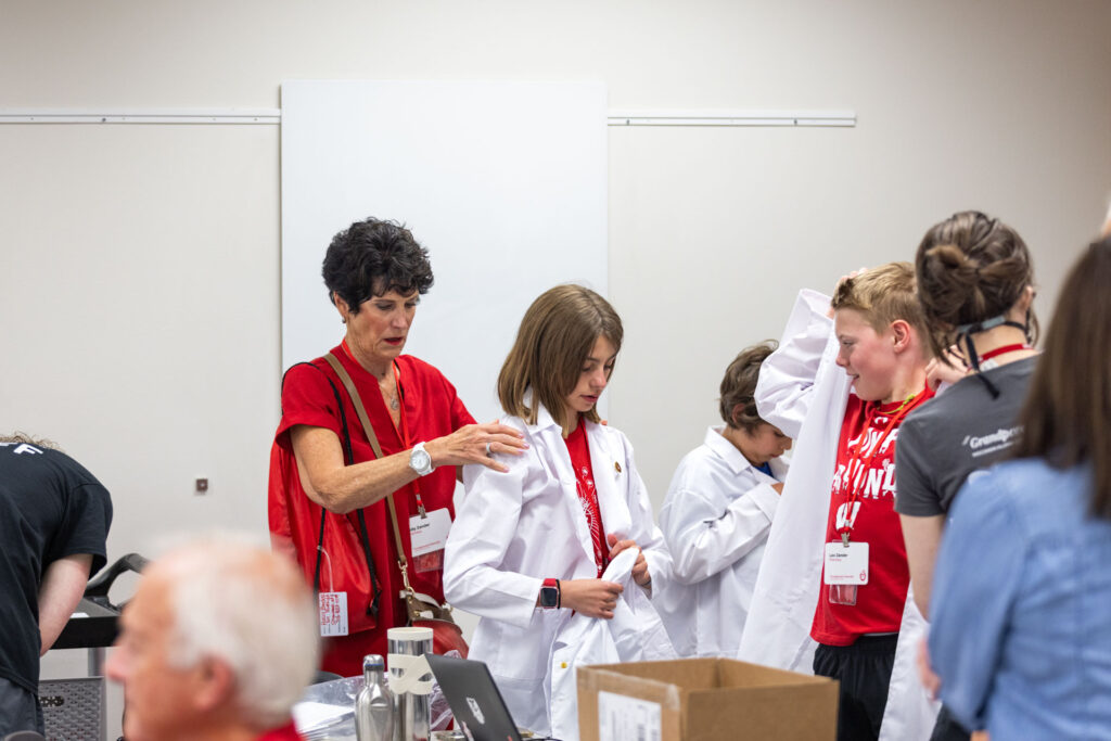An older woman in a red shirt helps a young girl put on a white lab coat. Other children are also putting on lab coats in the crowded room.