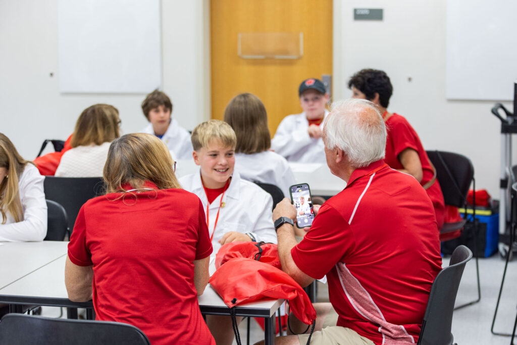 A seated older man in a red shirt shows a photo on his phone to a smiling young boy in a white lab coat, who is seated across the table. An older woman in a red shirt is seated in the foreground with her back to the camera.