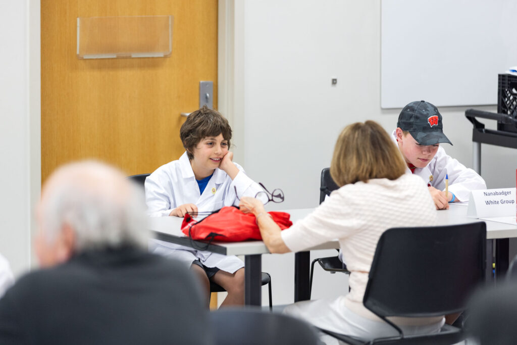 A boy in a white lab coat and a young man in a white lab coat and black baseball cap are seated at a table. The boy is smiling and leaning his head on his hand as a woman seated across from him passes him a pair of glasses.