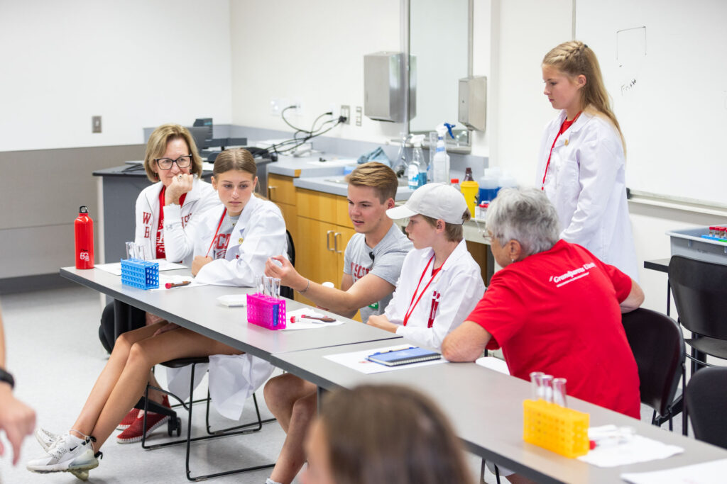 A group of participants, including two older women and three young people in white lab coats, are seated at a long table during a lab session. One girl in a lab coat stands behind the table, watching the others.