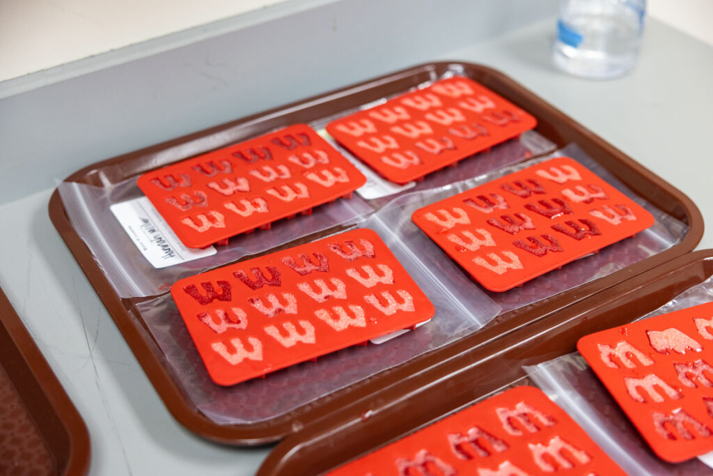 An overhead shot of several brown trays holding multiple red silicone molds, each filled with a partially set red, gelatinous substance in the shape of the letter "W."