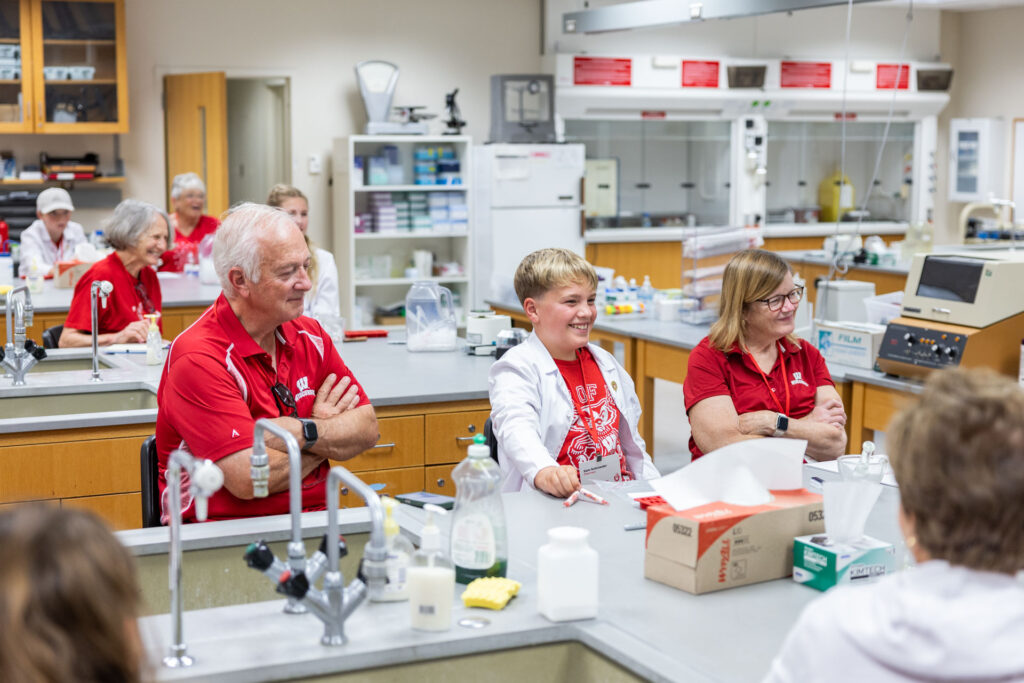 A group of participants in a bright lab, including two older adults in red shirts and a boy in a white lab coat, seated at a lab bench and smiling at someone off-camera.