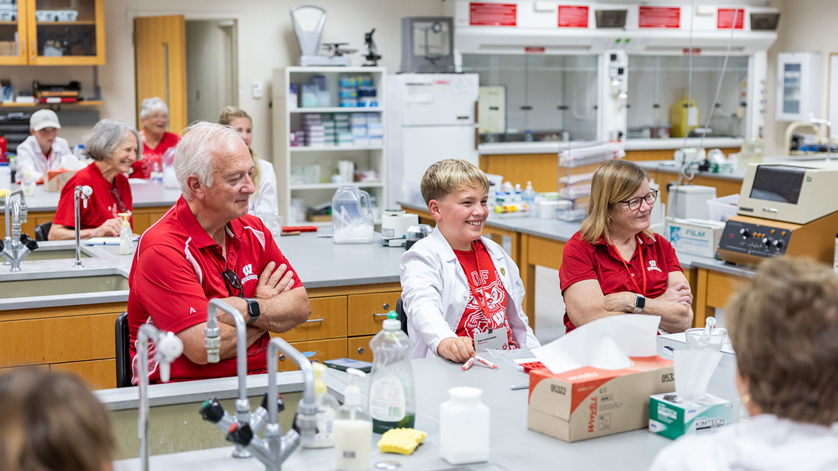 An older man sits next to a lab table with a young boy and another older woman. All wear Badger red, and the boy also dons a white coat.