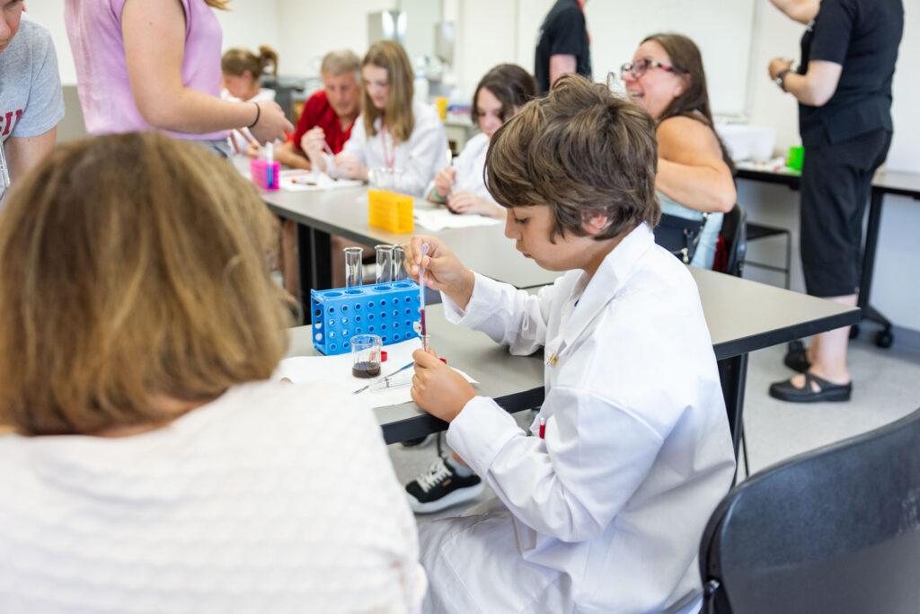 A young boy in a white lab coat is seated at a long table, focusing on using a dropper to transfer liquid into a test tube in a blue rack. Other adults and students are visible engaging in activities at the surrounding tables.