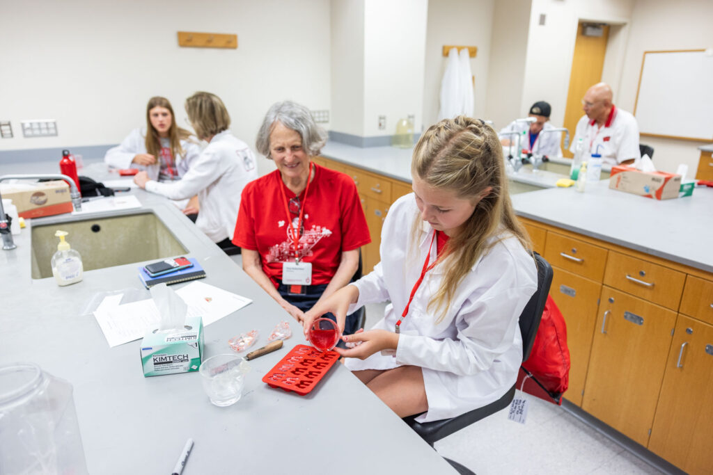 A girl in a white lab coat pours a red liquid from a small beaker into a red silicone mold, while an older woman in a red t-shirt sits across from her and smiles in the background. Other participants are visible in the lab.