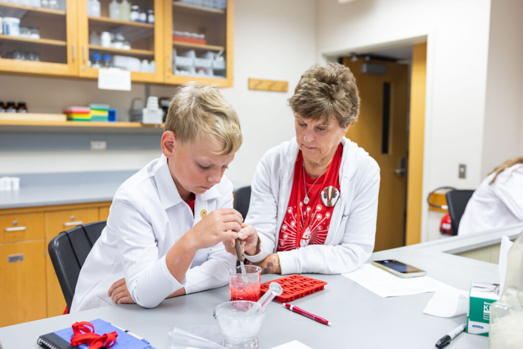 A young boy in a white lab coat and an older woman in a white sweater over a red shirt, sit at a lab bench and work together to mix a red substance in a small beaker or mortar.