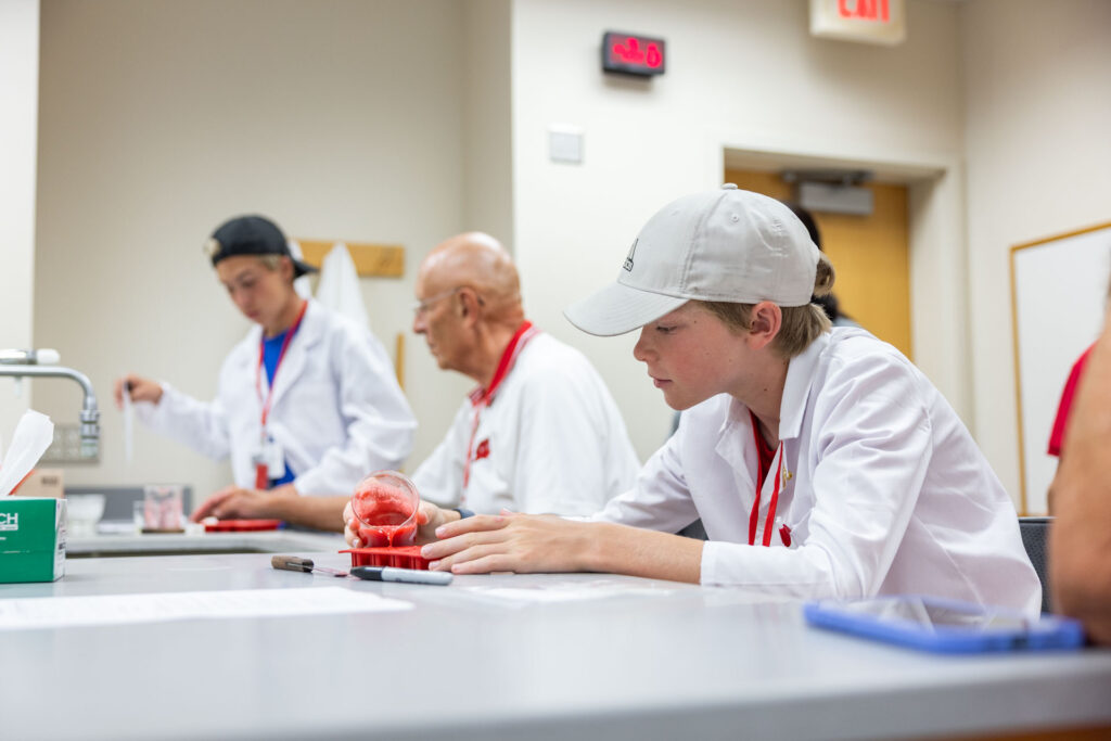 A young boy in a white lab coat and a gray baseball cap is focused on a small beaker of red liquid on a lab bench. An older man in a red shirt and another student are visible in the background.