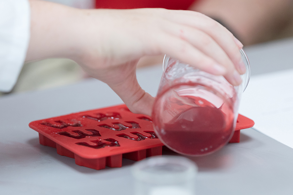 A hand pours a beaker of red liquid into a mold