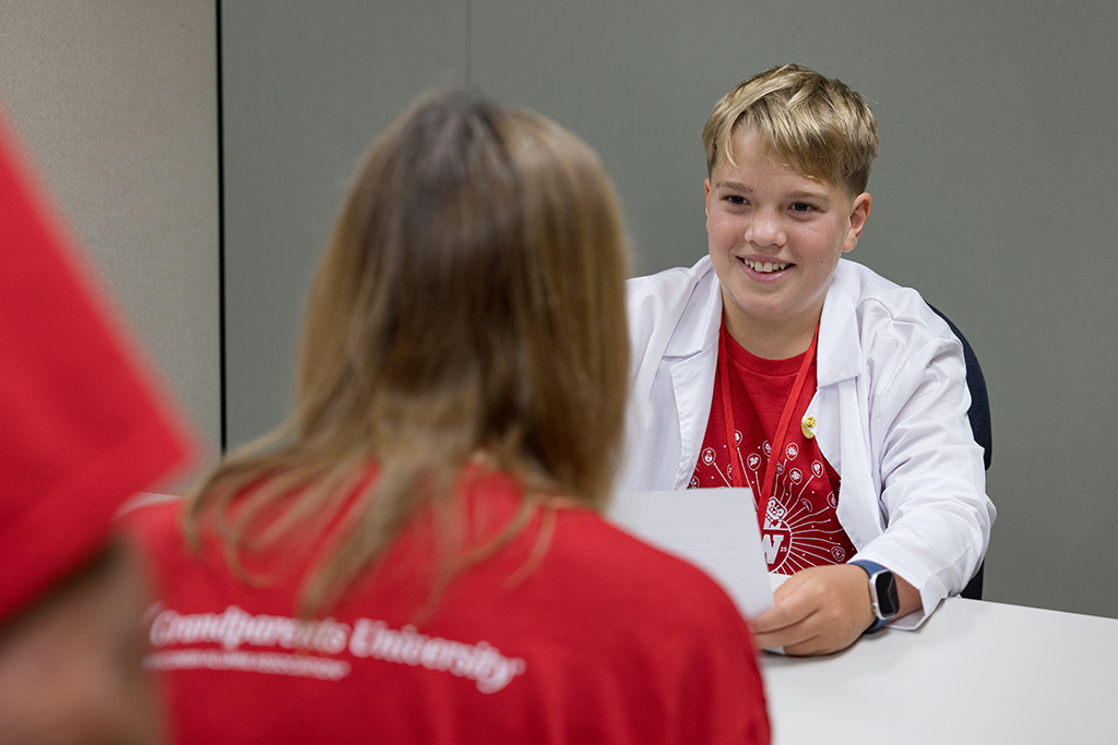 A close-up of a smiling young boy in a white lab coat and a red t-shirt, holding a sheet of paper while talking to an adult, who is seated opposite him and is only visible from the back. The adult's red shirt has the text "Grandparents University" visible across the shoulders.